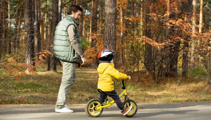 Young man walking by his little son in safety helmet and casualwear riding balance bicycle along wide road in park while enjoying chill at leisure